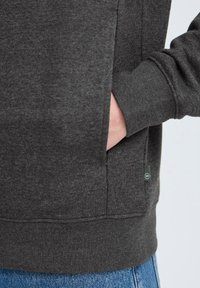 Grå sweatshirt med en myk tekstur; har en sidelomme og grønn logoetikett nær kanten; uformell, avslappet passform synlig.