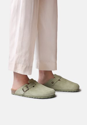 Osoba nosząca jasnobeżowe spodnie skrócone i zielone zamszowe chodaki na ślizg z regulowanymi paskami na klamrę na jednolitym tle.