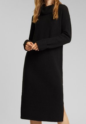 Femme portant une longue robe-pull noire en maille avec un col montant et des poignets côtelés, debout devant un fond gris uni.