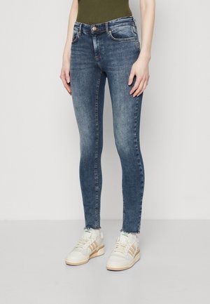 Skinny blue jeans with a faded finish, five-pocket design, and frayed hem. Worn with white and beige sneakers.