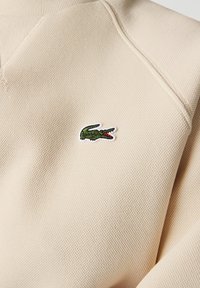 Sudadera de punto beige con cuello de canalé, que presenta un logo bordado de cocodrilo verde con acentos rojos en el pecho. Tejido texturizado.