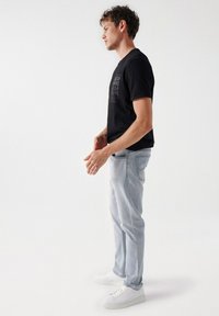 Camiseta negra de manga corta con una marca texturizada, combinada con jeans azul claro y zapatillas blancas, mostrada de lado sobre un fondo liso.