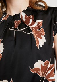 Sort bluse med blomsterdesign, der har cremede og brune blomstermønstre, plisseret halsudskæring og korte ærmer med samlet detalje.
