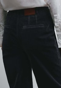 Zwarte corduroy broek met een gestructureerde afwerking, voorzien van achterzakken en een bruin leren label in de taille, gecombineerd met een wit overhemd.