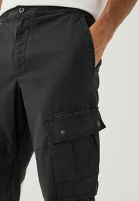 Black cargo pants made of lightweight fabric, featuring multiple pockets, snap buttons, and a relaxed fit at the waist. Textured finish visible.