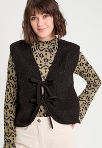 Gilet di pile nero con scollo a V, legature frontali e rifiniture nere a contrasto, indossato sopra una maglietta a fantasia verde e abbinato a pantaloni di colore chiaro.