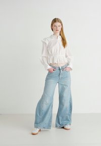 Blusa blanca a rayas con acentos con volantes, combinada con pantalones vaqueros de pierna ancha en azul claro. La modelo lleva zapatillas blancas, frente a un fondo liso.