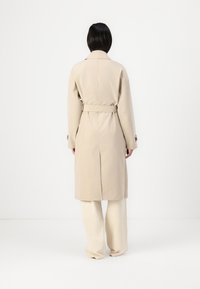 Beige trench coat övvel a deréknál, hosszú ujjak, és oldalsó hasítékokkal. Simább textúrával, térd hosszúságú, krém színű, széles lábukra esik.