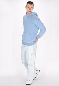 Felpa con cappuccio azzurro chiaro con tasca frontale, indossata con jeans cargo azzurro chiaro dotati di tasche laterali, abbinata a sneaker bianche.