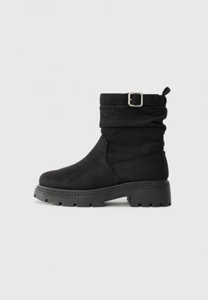 Bottine noire avec un design flou, fabriquée en matériau doux, dotée d'une boucle sur le côté et d'une semelle épaisse à crampons pour une bonne adhérence.