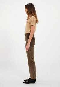 T-shirt beige à manches courtes, pantalons moulants verts olive, silhouette effilée, tissu lisse, chaussures noires avec détails de logo. Fond neutre.