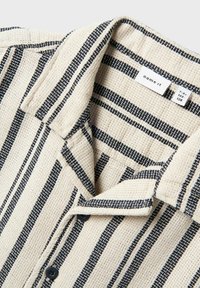 Camicia a maniche corte a righe beige e blu navy, realizzata in tessuto testurizzato. Presenta un colletto e bottoni per la chiusura. Etichetta del marchio visibile all'interno.