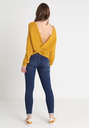 Femme portant un jean skinny bleu foncé, un pull dos nu jaune moutarde avec un nœud et des baskets beige, debout de dos sur fond blanc.