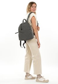 Junge Frau trägt cremefarbene Hose, weißes ärmelloses Oberteil, beige Sneaker, mit einem grauen gesteppte Rucksack über der Schulter, steht vor weißem Hintergrund.