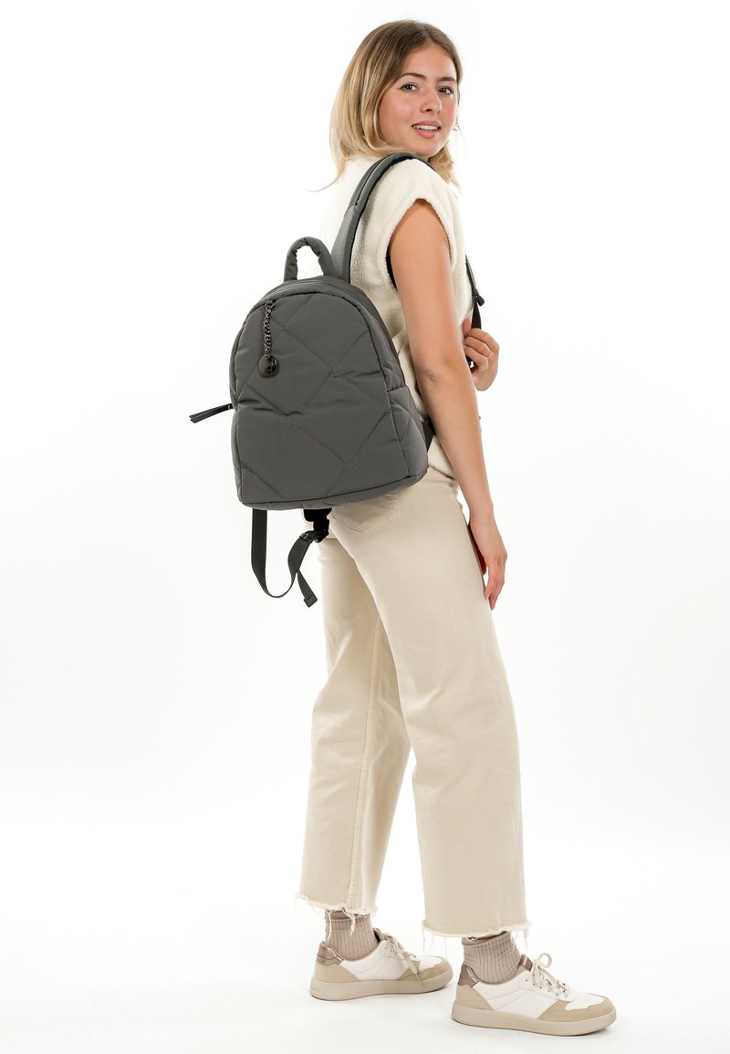 Junge Frau trägt cremefarbene Hose, weißes ärmelloses Oberteil, beige Sneaker, mit einem grauen gesteppte Rucksack über der Schulter, steht vor weißem Hintergrund.