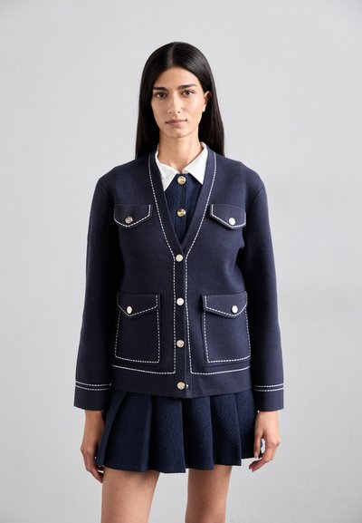 Cardigan bleu marine avec des coutures blanches, deux poches avant, et des détails de boutons dorés, porté sur une jupe plissée assortie.