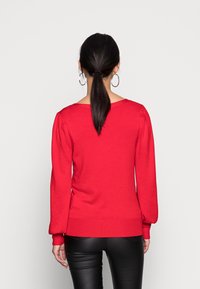 Pull en maille rouge avec un large décolleté et des manches longues bouffantes, doté de poignets et d'un ourlet côtelés. Associé à un pantalon noir ajusté.