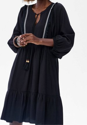 Schwarzes Kleid mit langen Puffärmeln, V-Ausschnitt, gesticktem weißen Besatz und Quasten-Details. Zartes Material mit einem gestuften, ausgestellten Saum.