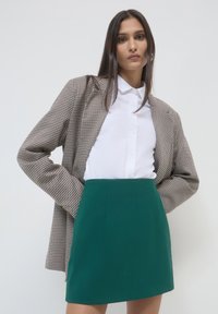 Blazer brown in geometria houndstooth sopra una camicia bianca con bottoni, abbinato a una gonna mini verde a tinta unita. Il tessuto liscio e la sartoria elegante sono evidenti.