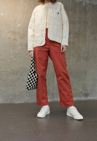 Prošívaná krémová bunda, červené kalhoty, bílé tenisky a černobílá kostkovaná taška tvoří ležérní outfit na texturovaném šedém pozadí.