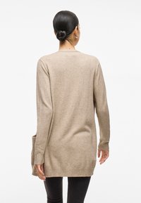 Long beige cardigan made of soft knit material. Features ribbed cuffs and hem, with a loose fit and side pockets. Worn over black pants.