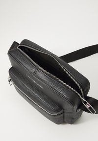 Texturiertes schwarze Lederhandtasche mit zwei Reißverschlussfächern und einem verstellbaren Tragegurt. Ausgestattet mit silbernem Zubehör und einem eingeprägten Logo auf der Vorderseite.