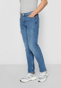 Homme portant un jean bleu slim et des baskets blanc-gris, debout avec une main dans la poche devant un fond uni.