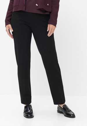 Persona che indossa pantaloni neri a gamba dritta, mocassini in pelle neri e una giacca bordeaux scura con bottoni dorati, in piedi su sfondo bianco.
