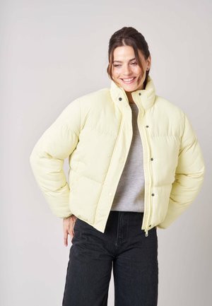 Vrouw die glimlacht, draagt een lichtgele donsjas, een grijze trui en zwarte broek, staat met één hand op haar heup tegen een effen achtergrond.