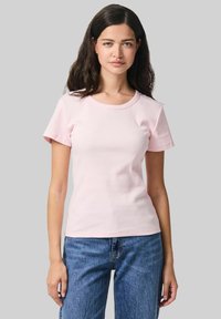 Gaiši rozā, īsām piedurknēm, t-krekls, kas izgatavots no ribota auduma, ar apaļu izgriezumu. Apvienots ar ziliem džinsiem, modelis stāv pret pelēku fona.