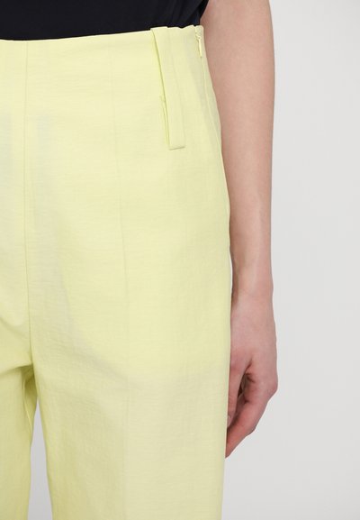 Pantalons à larges jambes de couleur jaune clair avec une texture lisse, comportant des poches latérales et une fermeture éclair, présentés de la taille jusqu'à mi-cuisse.