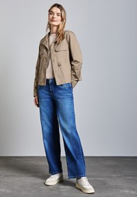 Ung kvinna som står upp, iförd en beige jacka, ljus tröja, vida blå jeans och vita sneakers mot en enkel grå bakgrund.