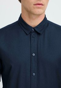 Camisa azul marino, de botones, con una textura suave y collar clásico. Presenta dos botones en el collar y un diseño limpio y minimalista.