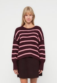 Pull en tricot avec de larges rayures bordeaux et rose, une coupe décontractée et des poignets côtelés. Associé à une jupe bordeaux foncé.
