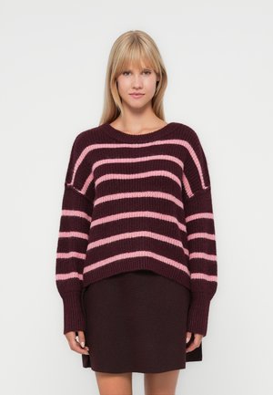 Strikkegenser med brede maroon og rosa striper, avslappet passform og ribbestrikkede mansjetter. Kombinert med en mørk burgunder skjørt.