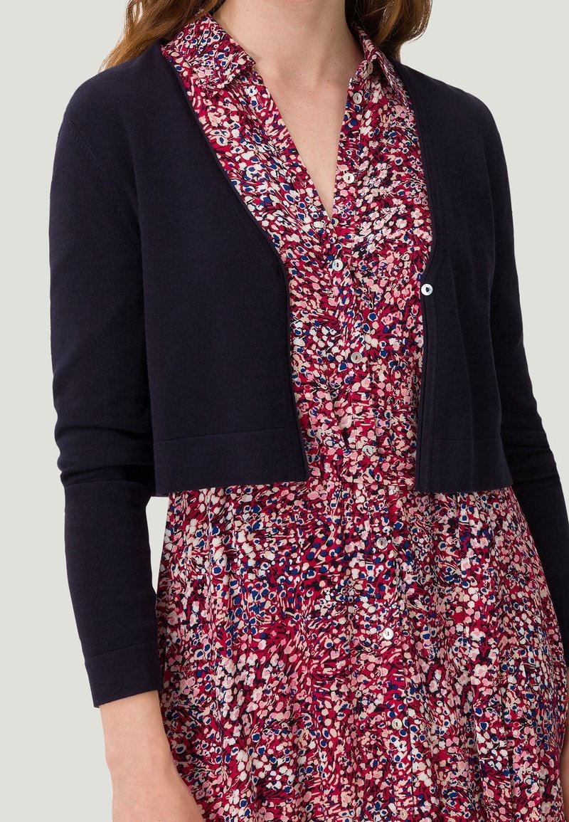 Femme portant un cardigan noir court par-dessus une robe rouge fleurie boutonnée avec des motifs roses, blancs et bleus, manches relevées jusqu'au poignet.