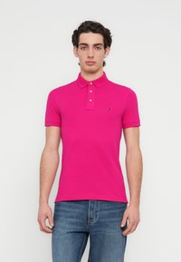 Ροζ εφαρμοστή πόλο μπλούζα από βαμβάκι με πλάκα τριών κουμπιών, κοντά μανίκια και μικρό λογότυπο στο στήθος. Φορεμένη με τζιν παντελόνι.