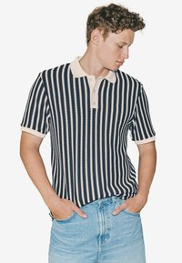 Polo shirt în dungi bleu marin și crem, cu guler, mâneci scurte și un model texturat, asortat cu blugi deschiși la culoare.