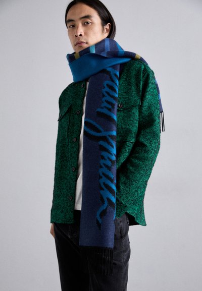 Eșarfă albastră și bleumarin cu un model decorativ și margini franjurate, purtată peste o jachetă verde cu un model. Contrast cu pantalonii negri.