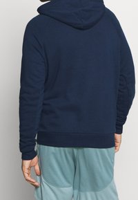 Felpa blu navy con cappuccio, polsini e fondo a coste. Presenta un tessuto morbido e una vestibilità rilassata, abbinata a pantaloni corti in rete color turchese chiaro.