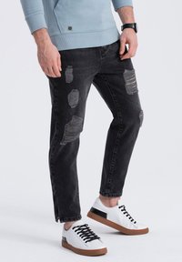 Vaqueros de mezclilla negros con detalles desgastados y dobladillos sin rematar, combinados con zapatillas blancas con acentos negros y suelas marrones.