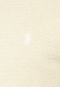 Krämfärgad stickad tröja med ett texturerat zigzagmönster och en liten, vit broderad logotyp på bröstet.