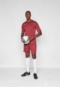 Hombre en uniforme de fútbol color burdeos sosteniendo un balón de fútbol en blanco y negro, vistiendo medias blancas hasta la rodilla y botas negras, de pie contra un fondo liso.