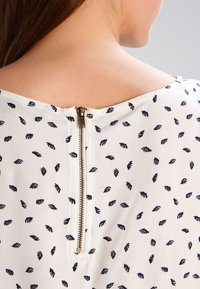 Abito in tessuto bianco con design a foglie blu; presenta una cerniera sul retro ben visibile e una scollatura ampia. Tessuto morbido e leggero.