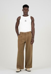 Pantalones marrones de pierna ancha con pliegues, combinados con una camiseta sin mangas blanca con un pequeño logo. El calzado incluye zapatos color crema con detalles.