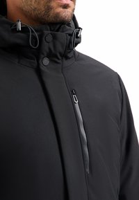 Man in een zwarte hoge kraagjas met knoop- en ritsdetails, en verstelbare koordsluitingen op de capuchon.