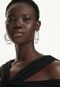 Silberne Creolen mit geschichtetem Design und strukturierten Oberflächen, getragen mit einem schwarzen drapierten Oberteil, das aus weichem Stoff und vertikalen Falten besteht.