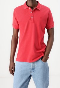Hombre vestido con una camiseta polo roja de manga corta con botones y jeans de mezclilla azul claros, de pie con una mano en el bolsillo contra un fondo blanco.