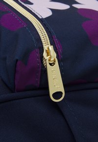 Laukkua navy-kangasta, jossa on kultaketju ja logotagi. Suunnittelussa on etupuolella violetteja ja vaaleanpunaisia abstrakteja muotoja.