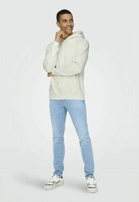 Young man smiling, wearing a light beige hoodie, light blue jeans, and white sneakers, standing against a plain background.